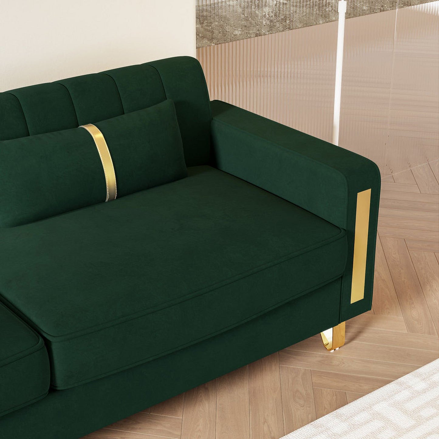 The Mid Century Modern Sofa Couch, upholstered in modern green velvet and featuring gold accents on the armrest and legs, sits on a wooden herringbone floor. A coordinating green cushion adorned with a gold stripe is placed on the sofa. In the background, part of a white wall and glass partition can be seen.