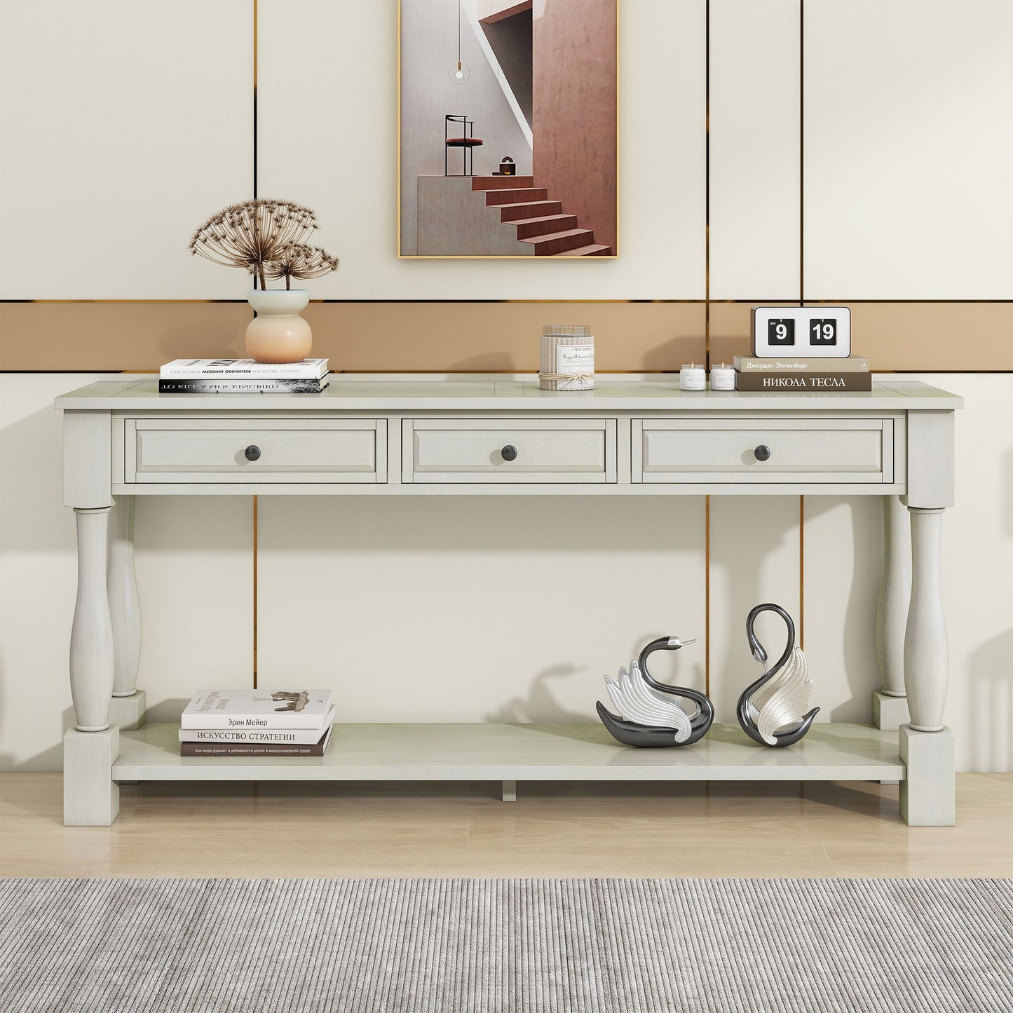 A light console table with three drawers and turned legs is adorned with a vase of dried flowers, a black and white flip clock, and a candle. Below, two swan figurines rest on stacked books. Modern wall art serves as the backdrop for this Console Table Long Console Table With Drawers And Shelf For Entryway, Hallway, Living Room.