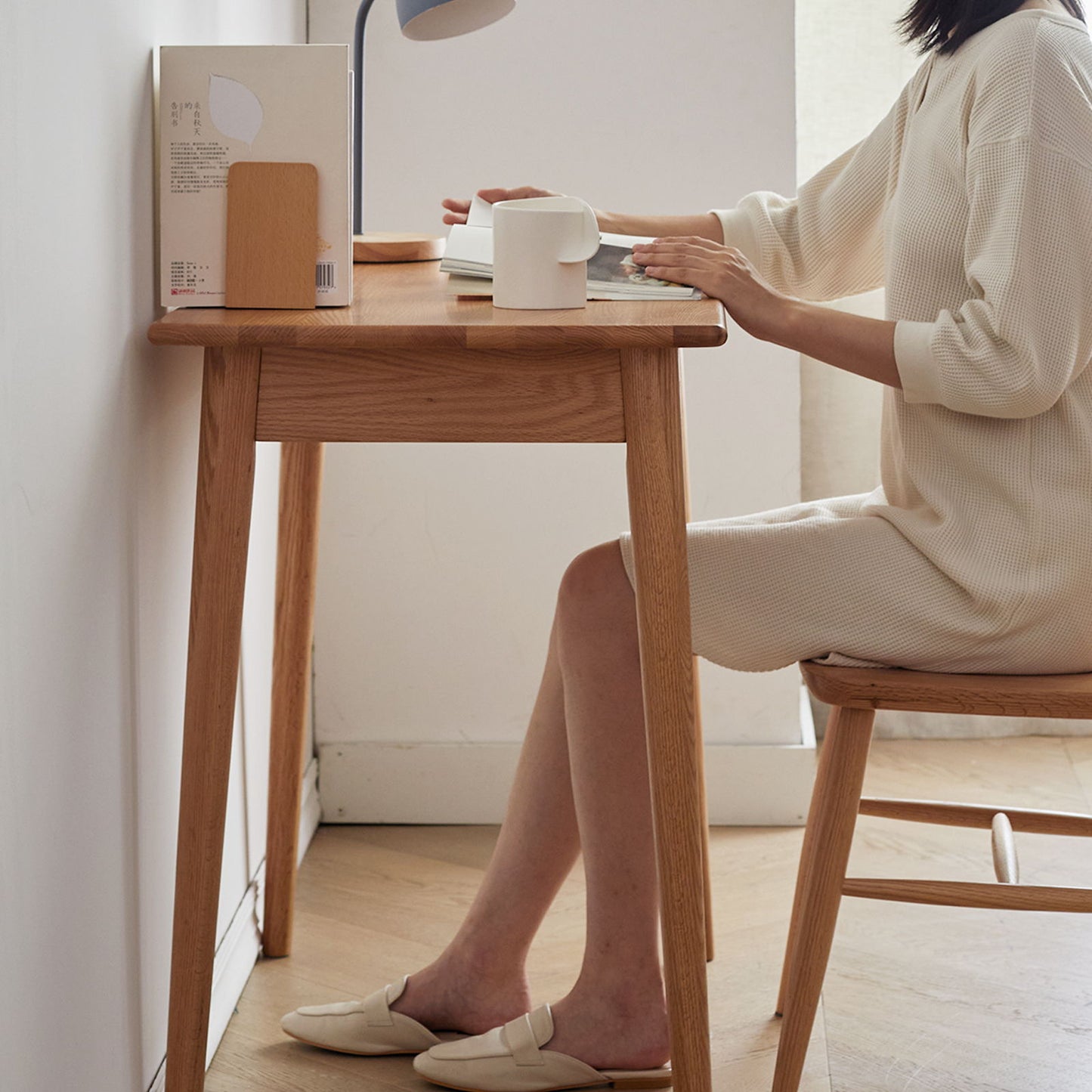 A person in a white sweater dress and beige slippers sits at an oak natural computer desk. The minimalist setting features books, a desk lamp, and a white mug under natural lighting.