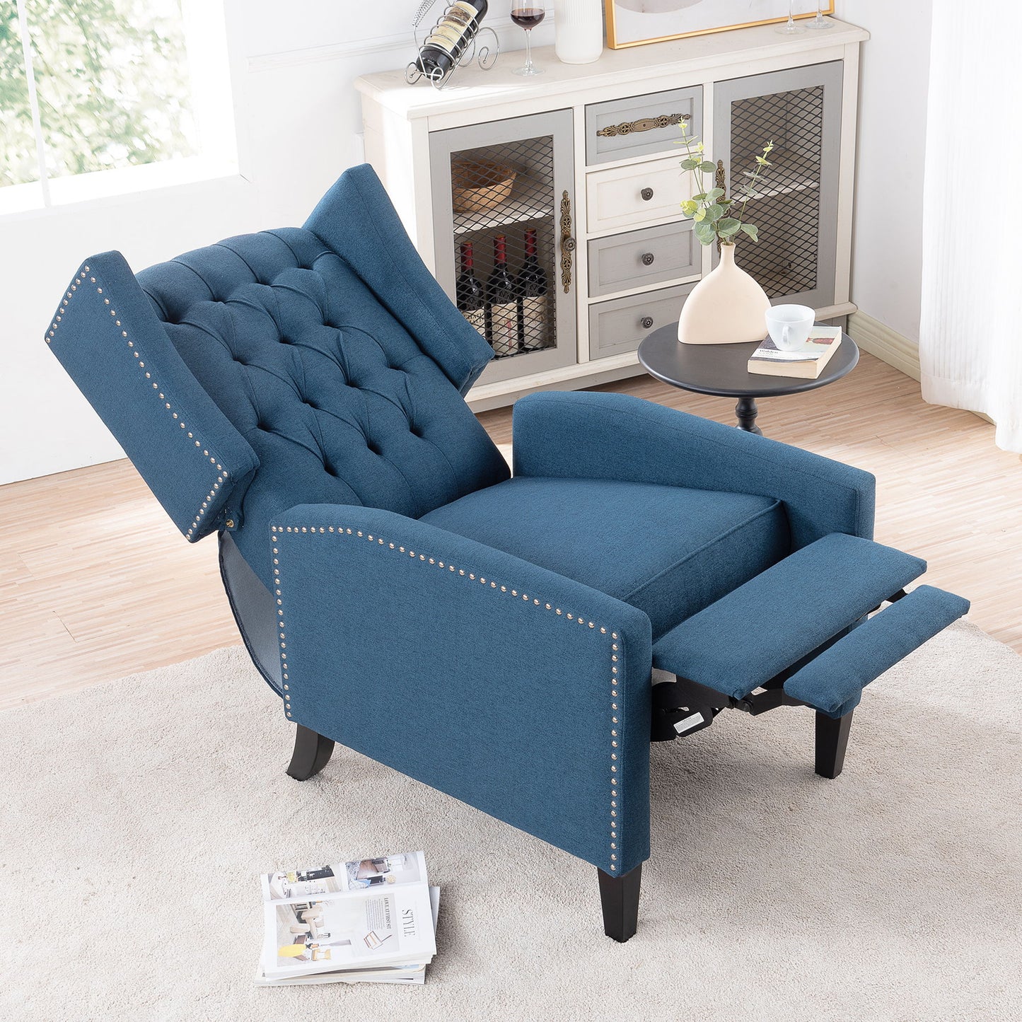 The Manual Wing Chair Recliner, tufted and with nailhead trim, sits in a cozy living room facing a round black side table holding a cup and book. Nearby is a cabinet featuring a wine rack and decor, set by a partially open window.