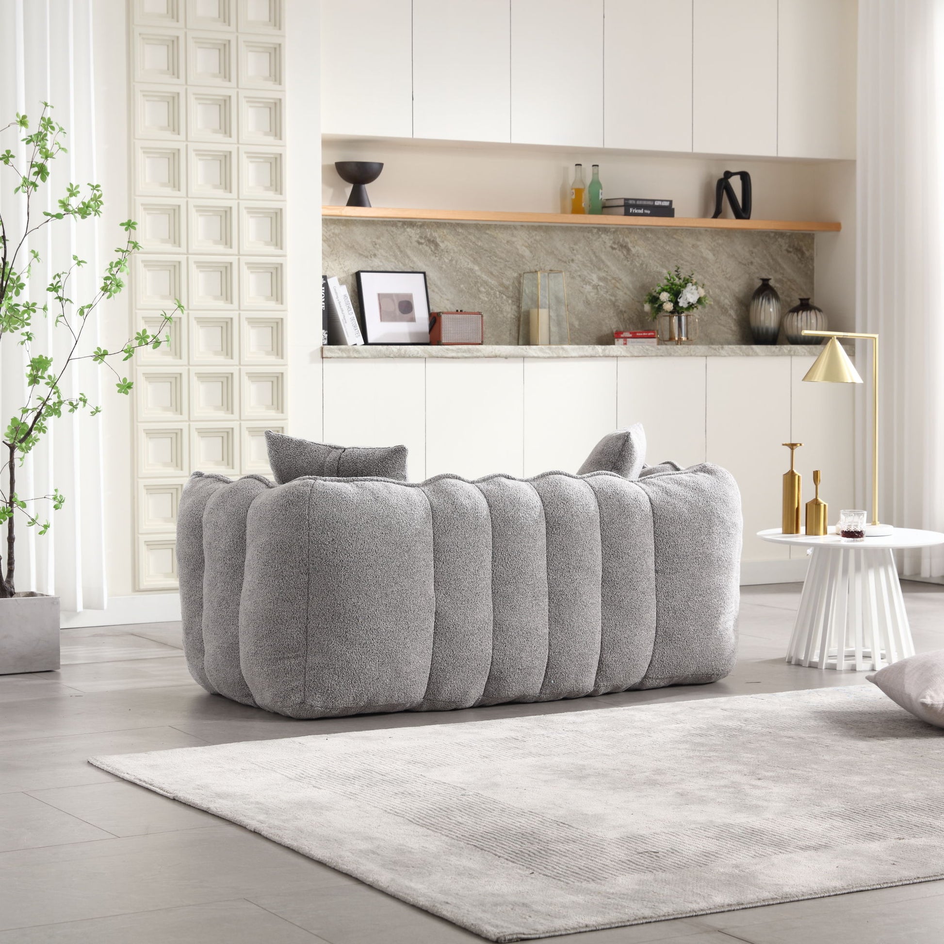 Modern living room with a plush gray sofa and matching rug. Behind the sofa, the kitchen area has white cabinets, marble backsplash, and a wooden shelf adorned with decor. A geometric lamp and small round table are on the right. A soft beanbag chair for two people adds extra seating.