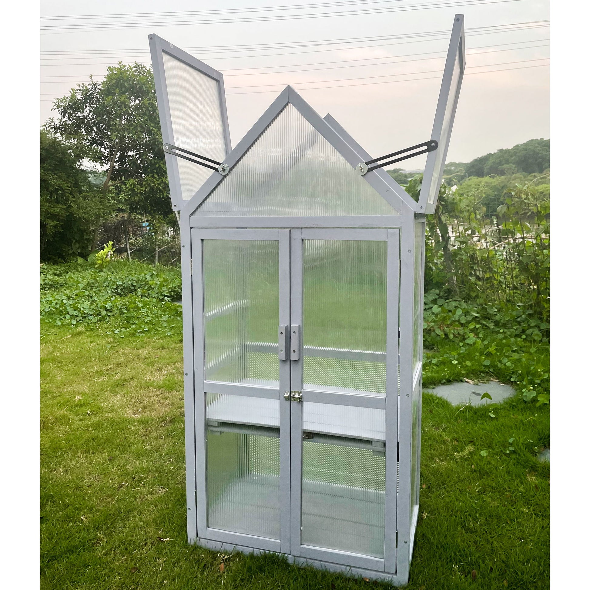 A gray, mini greenhouse with a peaked roof from the Mini Greenhouse Kit stands on grass. The top panels are open, surrounded by greenery and trees under a cloudy sky.