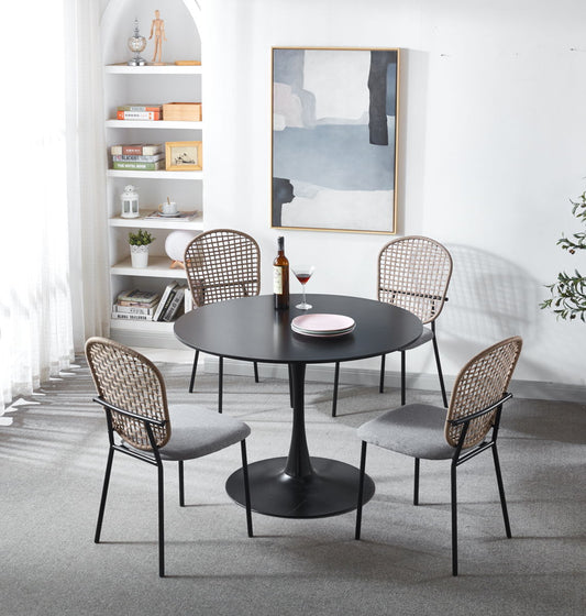 A modern dining room showcases a 5 Piece Dining Set in matt black and gray, complete with wine, a glass, and plates on a round table. Rattan-backed chairs surround it. A bookshelf, abstract wall art, and a plant enhance the decor as natural light flows through sheer curtains.