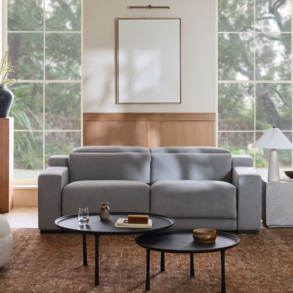 A contemporary living room showcases the Dalton Motion Sofa in light grey, set against a wooden wall panel. Above the sofa is a minimalist framed artwork. In front, two round black coffee tables are adorned with decorative items, and expansive windows provide a view of the lush greenery outside.