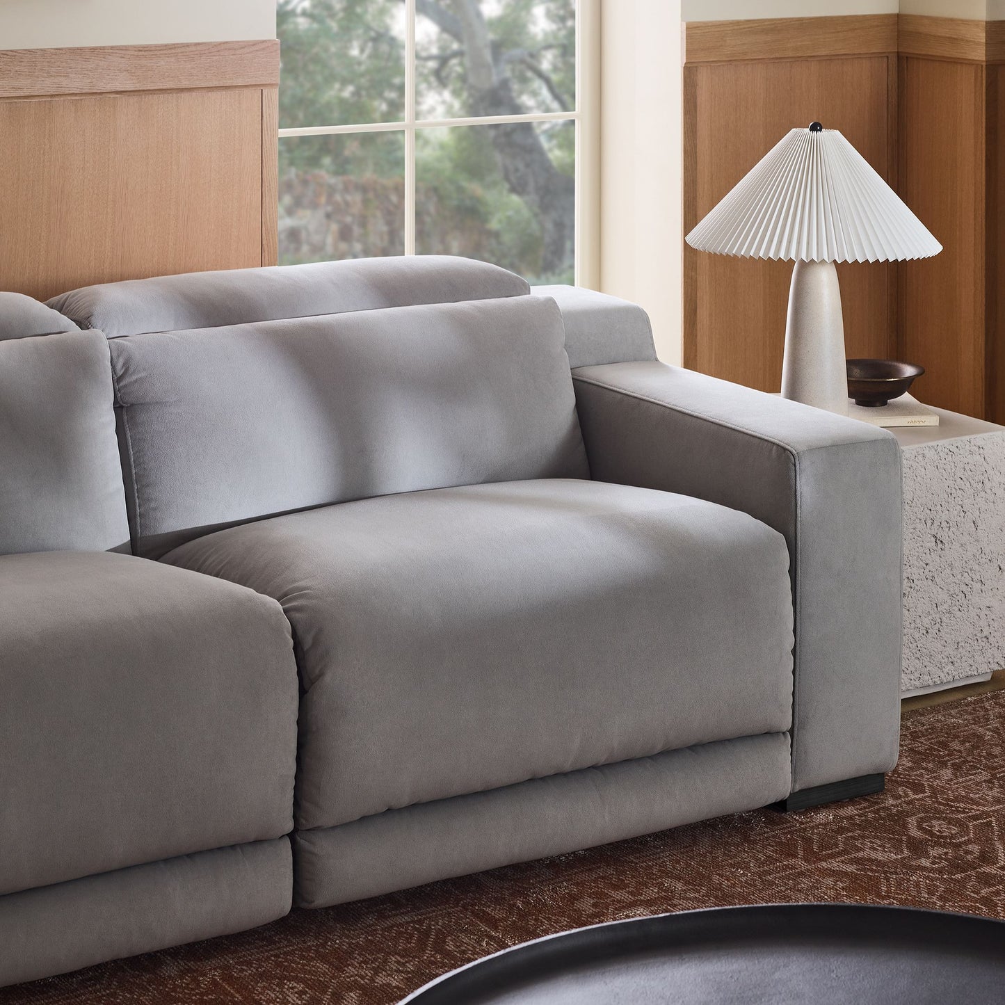 A modern living room featuring the Dalton Motion Sofa in a plush gray finish, complemented by a textured side table and a white pleated lamp. A large window offers an outdoor view, while wood paneling and a patterned rug complete the room's design.