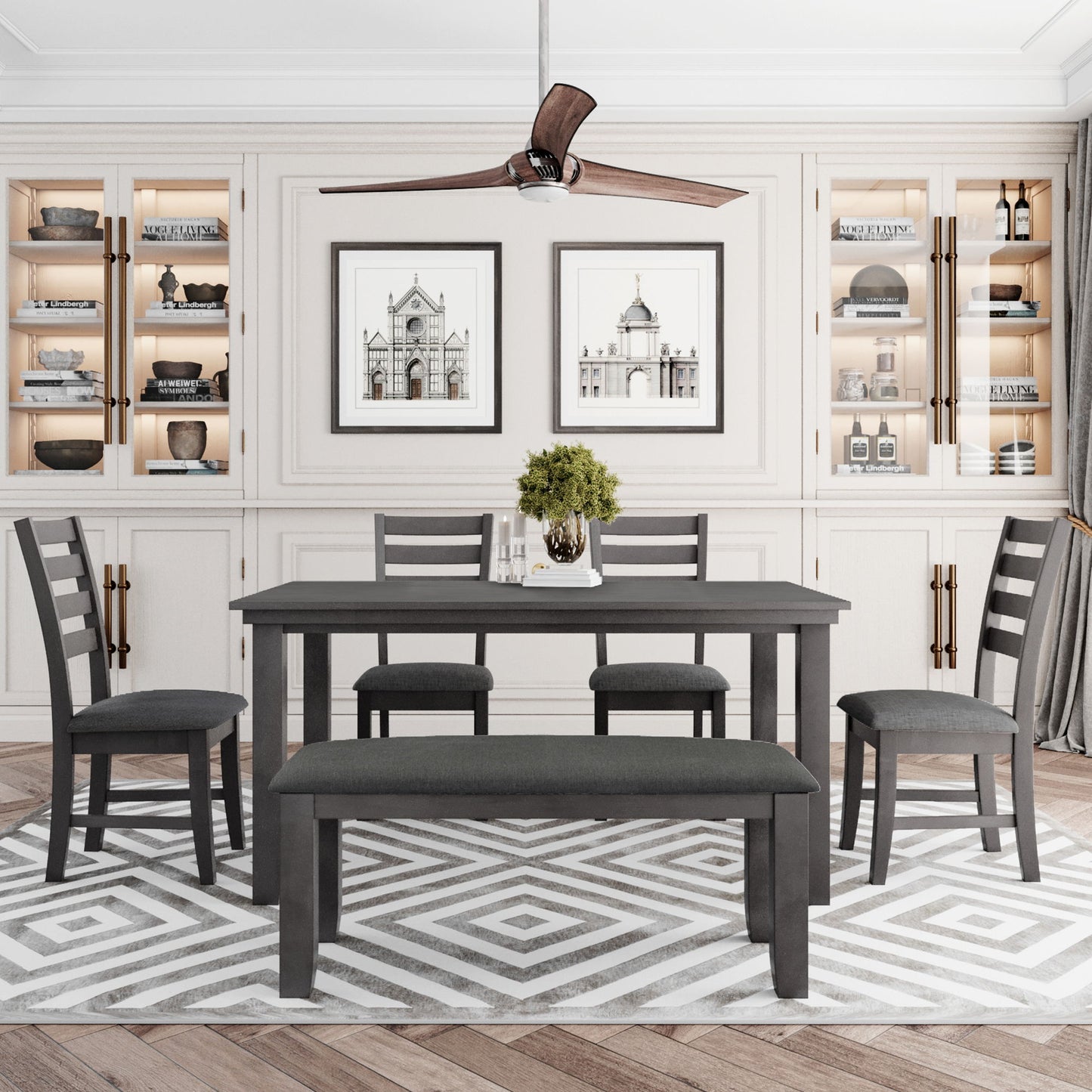 A modern dining room features a gray rustic wood dining set with a table, four chairs, and a bench on a geometric rug. The background has built-in shelves with decor and two architectural prints, while a ceiling fan hangs above.