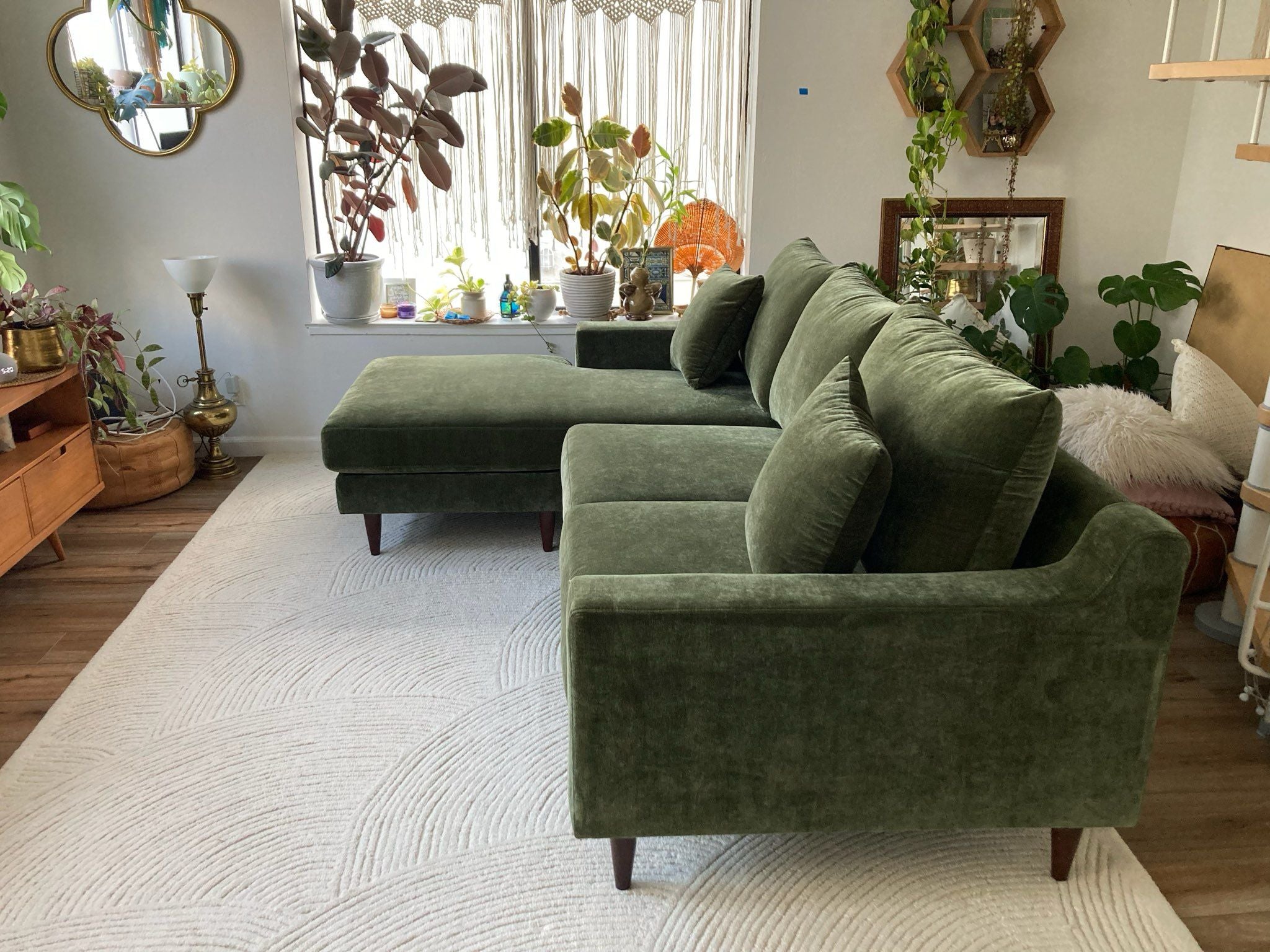 A sage green sofa, viewed from the side, on top of a textured white rug. Plants can be seen in the background. 