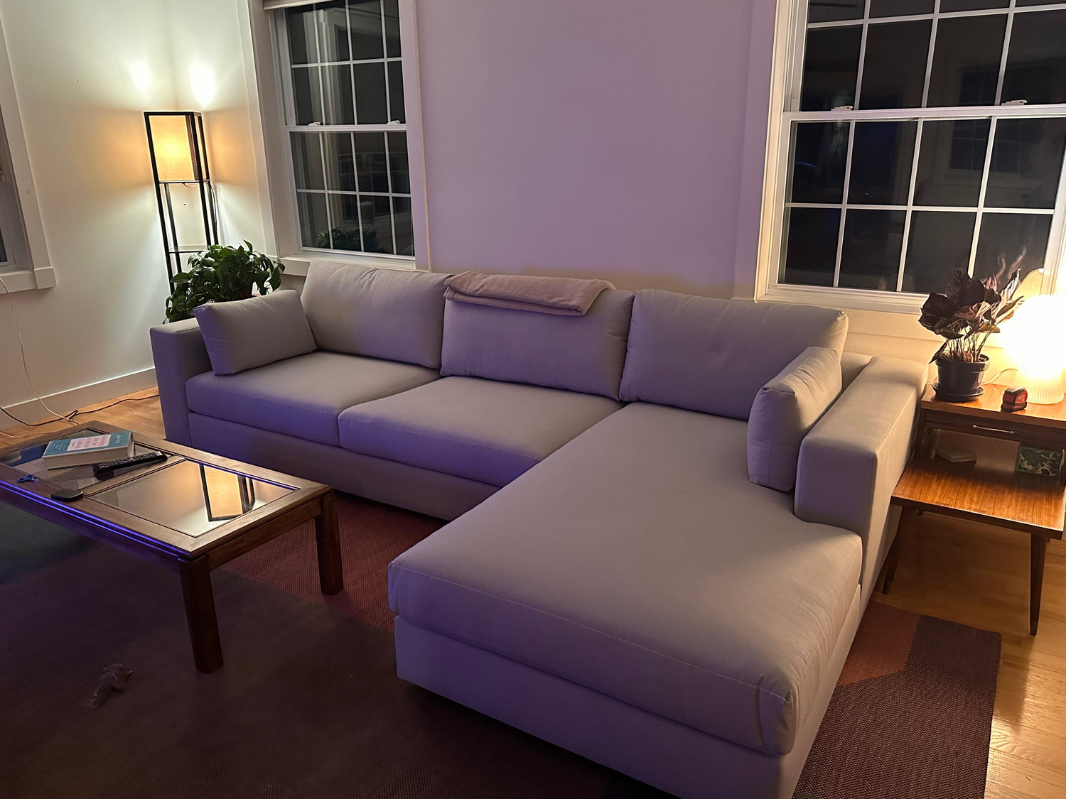 A cozy living room featuring a large L-shaped beige sofa, a coffee table with books, and a tall floor lamp. Soft lighting illuminates the space, alongside natural light from large windows. A small plant is placed beside the sofa.