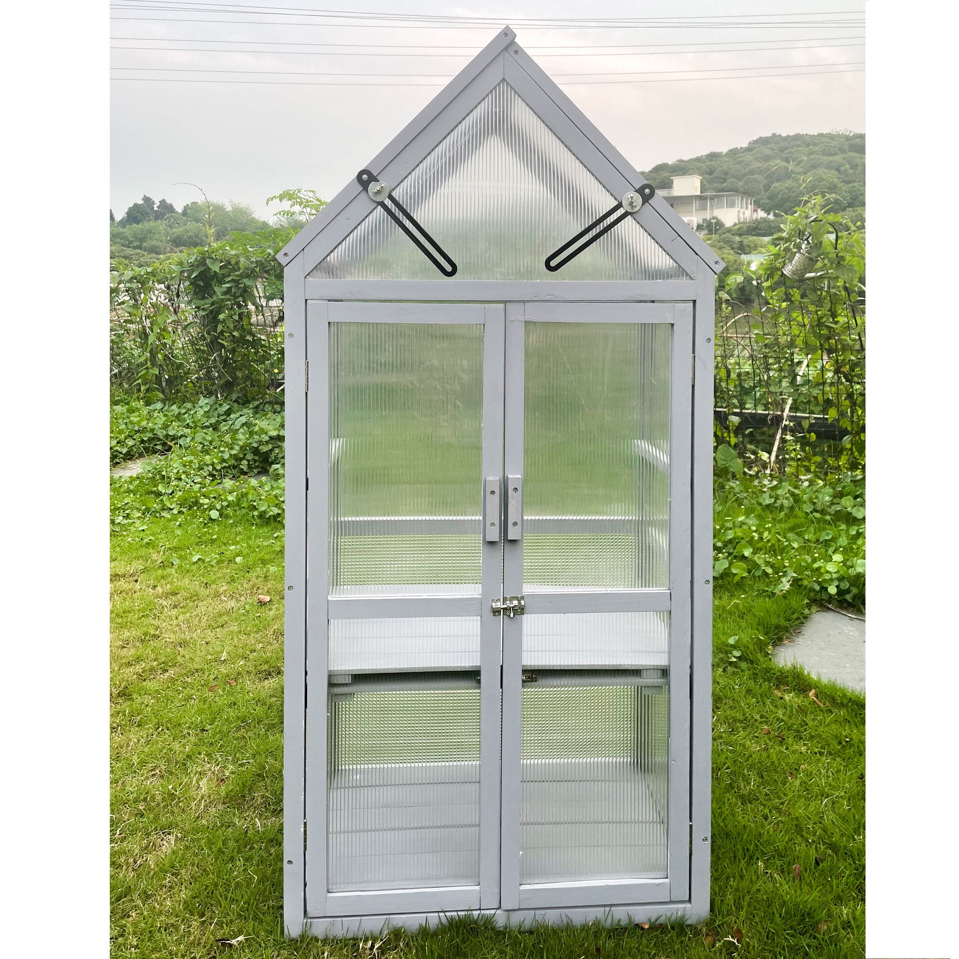 A Mini Greenhouse Kit with a peaked roof and transparent panels stands on the grass. It features a door with two horizontal handles and triangular top vents. Lush greenery and trees in the backdrop under an overcast sky complement its UV-resistant gray design, ideal for gardens, balconies, or patios.