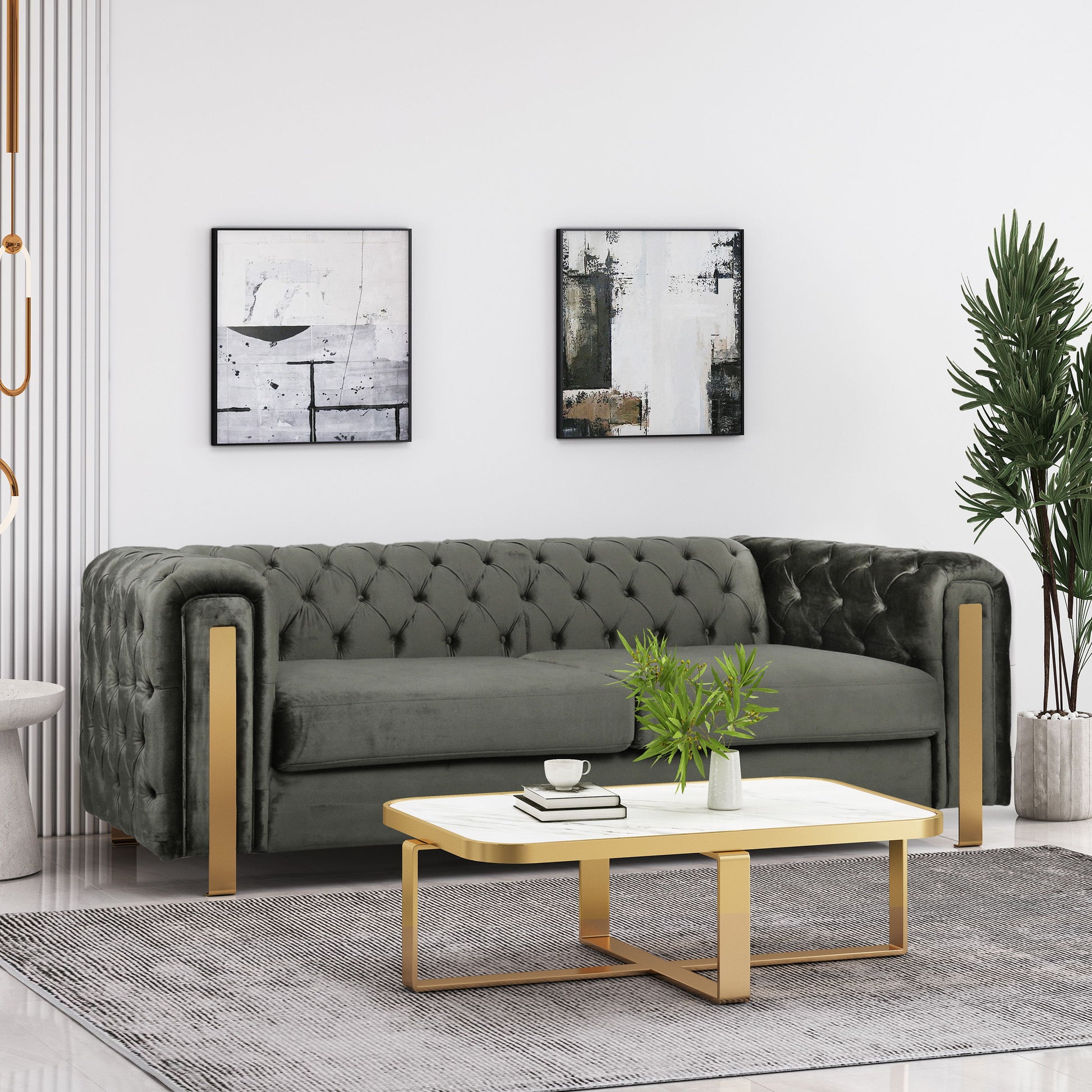 A modern living room showcases a gray Comfy 3 Sofa with tufted back and arms. Two abstract paintings hang overhead. A white rectangular coffee table with gold legs holds a plant and decorative book, while a tall potted plant is placed nearby on the light floor.