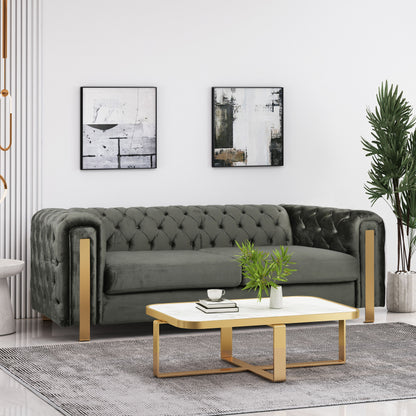 A modern living room showcases a gray Comfy 3 Sofa with tufted back and arms. Two abstract paintings hang overhead. A white rectangular coffee table with gold legs holds a plant and decorative book, while a tall potted plant is placed nearby on the light floor.