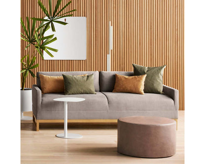 A modern living room features a gray sofa with green and brown pillows, a white round side table, and a brown round ottoman. A tall plant is on the left, and a mirror hangs on a wooden slat wall in the background.