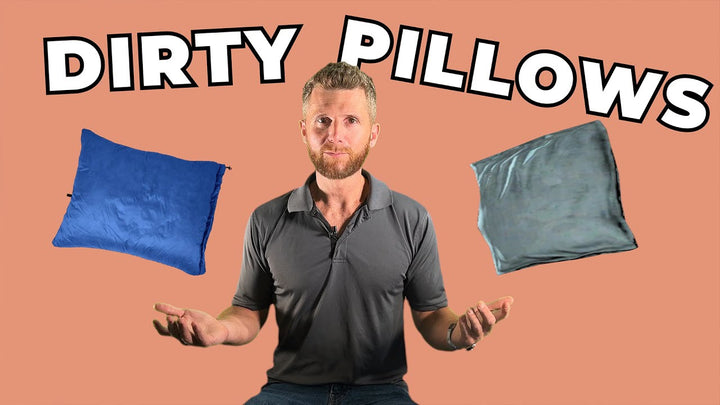 A man with a beard, wearing a gray shirt, stands against a peach-colored background with the words "DIRTY PILLOWS" at the top. He is holding out his hands, with a blue pillow on his left and a gray pillow on his right.