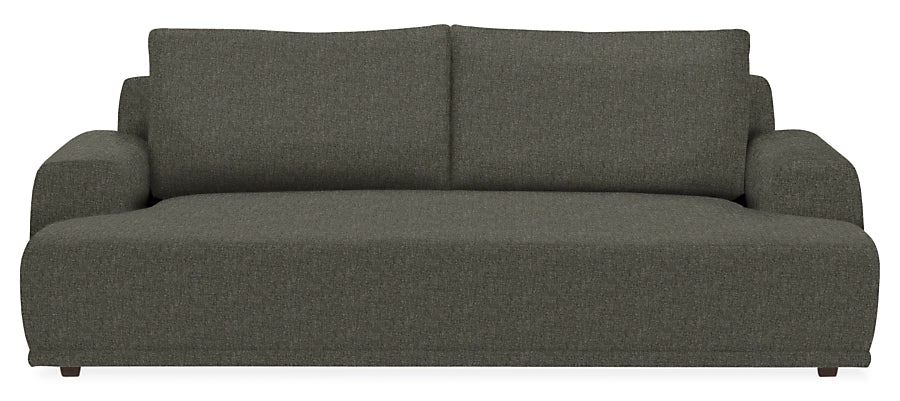 The Fia 90" Sofa is a large, modern dark gray piece with a low-profile design. It features thick cushions and wide armrests, set against a plain white background to highlight its minimalist style and comfortable appearance.