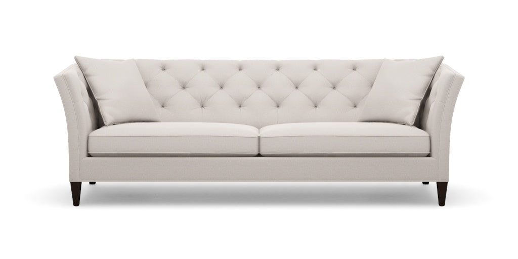 Introducing the Shelton 98" Sofa: a cream-colored masterpiece with a contemporary flair, featuring a tufted backrest and two perfectly matching throw pillows. The design boasts sleek, slightly flared arms complemented by elegant wooden legs, all showcased against a plain white backdrop.