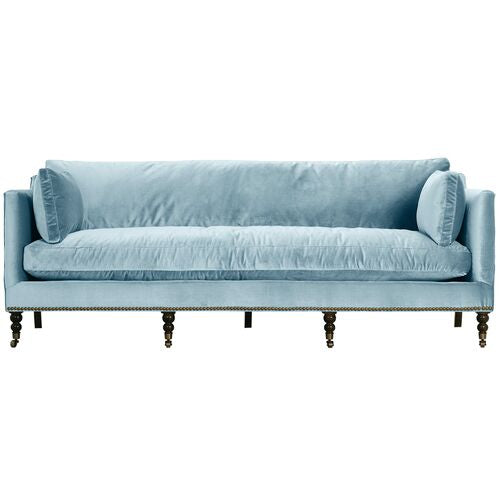 Introducing the Margot Velvet Sofa: a light blue sofa featuring a luxurious velvet fabric, complemented by a spacious long cushioned seat and cylindrical side cushions. It boasts a sleek straight backrest and stands gracefully on dark wooden legs with subtle casters for easy mobility.