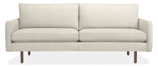 The 86" Jasper Sofa is a modern piece with a minimalist design, characterized by its light beige tone, sleek silhouette, and slim legs. It features two large cushions and appears upholstered in a soft fabric.