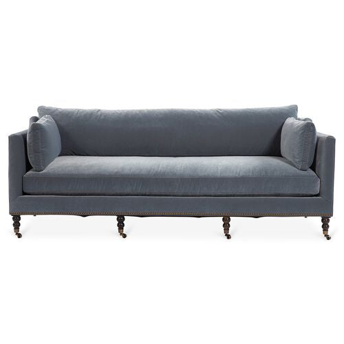 A Margot Velvet Sofa in blue, featuring two large cushions and cylindrical armrest pillows. The sofa is enhanced with wooden legs equipped with small wheels, creating a classic and elegant look.