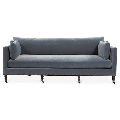 A Margot Velvet Sofa in blue, featuring two large cushions and cylindrical armrest pillows. The sofa is enhanced with wooden legs equipped with small wheels, creating a classic and elegant look.