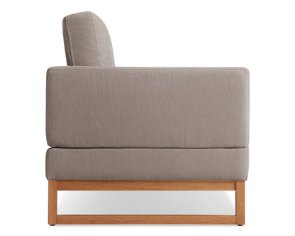 Side view of a modern gray upholstered armchair with a light wooden base. The chair has a rectangular shape and a minimalist design.