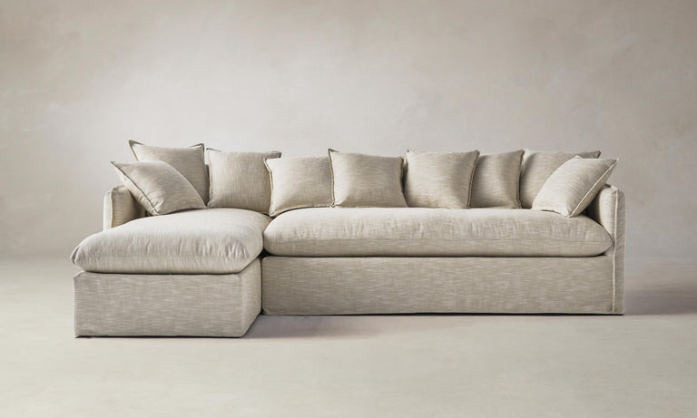 The Dune 90" Sectional in Belgian Linen Alder, a beige L-shaped sofa with multiple matching cushions, is set against a plain, light-colored wall. The left side includes a chaise lounge, complementing its minimalist and modern design.