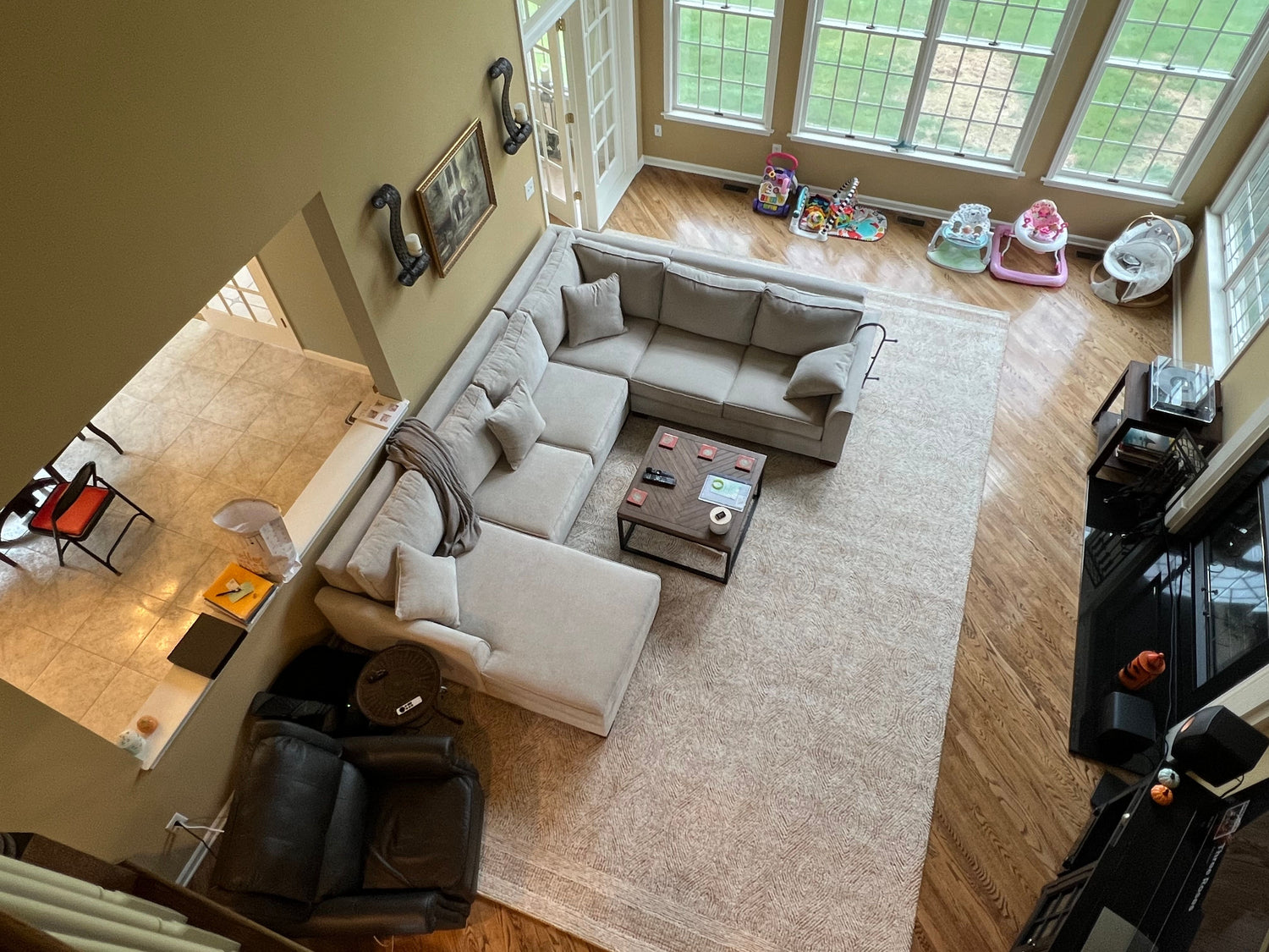 Aerial view of a living room with a large beige sectional sofa, coffee table with items, recliner chair, and baby toys by the windows. The room has hardwood floors and views of a tiled dining area.