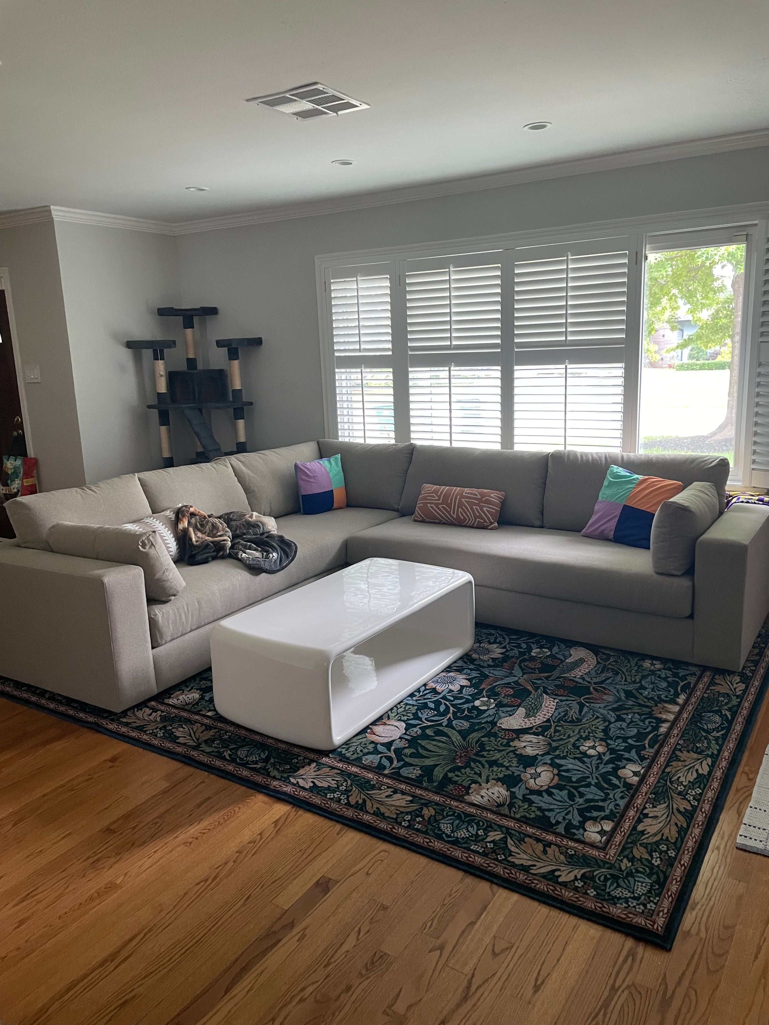 A cozy living room with a large gray L-shaped sofa featuring colorful pillows. There's a white coffee table on a patterned rug, a cat lounging on the sofa, and a tall cat tree in the corner. Sunlight streams through the window shutters.