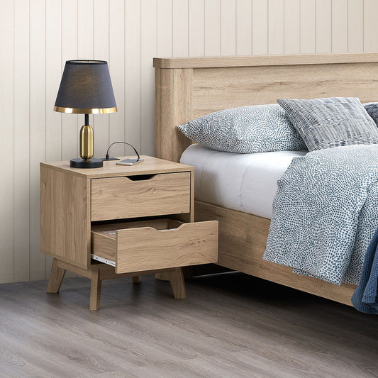 A Nordica nightstand in natural oak with USB/Type-C charging and pre-assembled drawer slides is beside a bed with a wooden headboard. It holds a black lamp and glasses, while the neatly made bed features a white duvet and patterned gray pillows against light wood paneling.