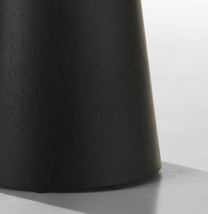 A close-up of the Jasper Round Table reveals its dark, textured, cylindrical form with a slightly tapered base against a smooth, light backdrop. The lighting accentuates subtle shadows on its surface, highlighting the matte finish.