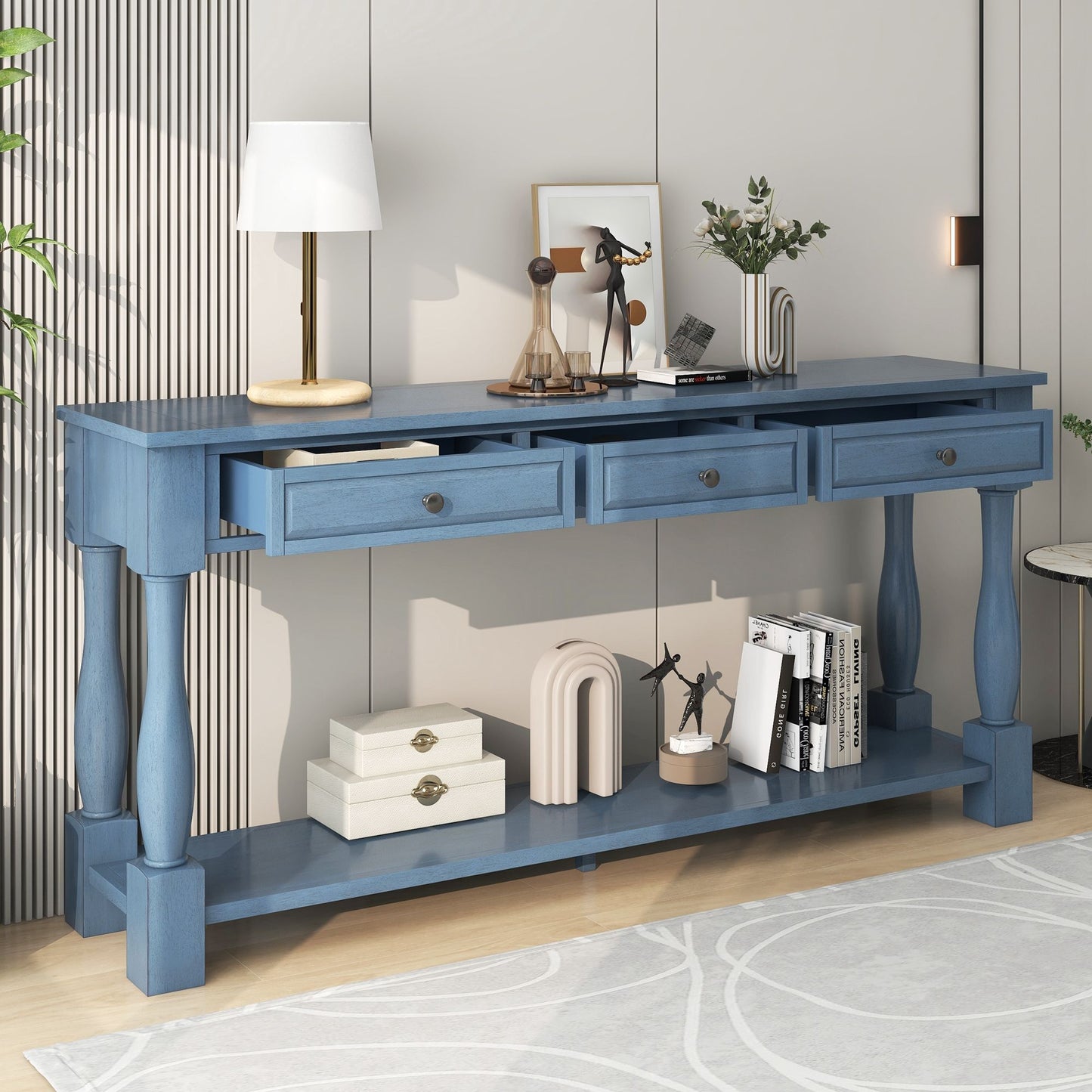 The Console Table Long Console Table with Drawers and Shelf in blue, featuring four drawers and turned legs, stands against a beige wall and holds a lamp, figurines, and a plant. Books and decorative boxes rest on the shelf below. A light gray rug covers the wooden floor.