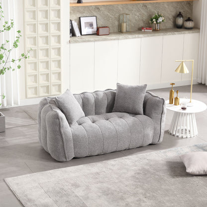 A cozy, modern living room showcases a soft beanbag chair with a high resilience foam core for two people, a white round table displaying a gold lamp and decor, and a textured rug. Natural light streams through the window, illuminating greenery and neutral wall art.