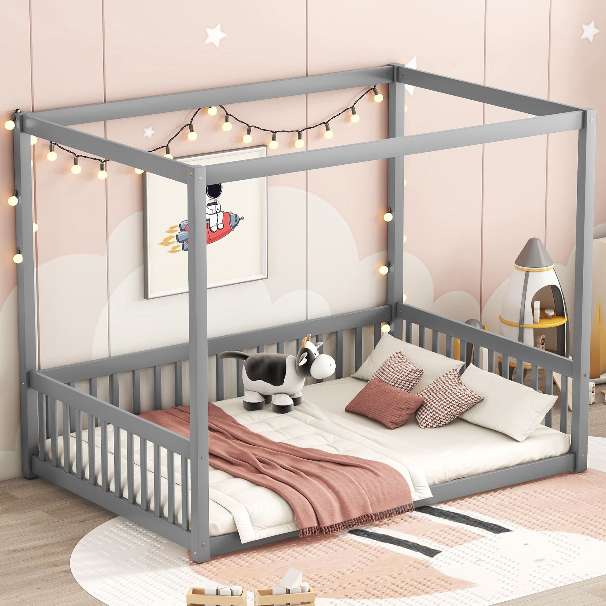 A children's bedroom featuring a Canopy Frame Floor Bed With Fence and Guardrails adorned with string lights. The soft pink walls have cloud and star decals. On the bed are a toy cow, striped pillows, and a pink blanket. In the corner, there's a small rocket-themed tent.