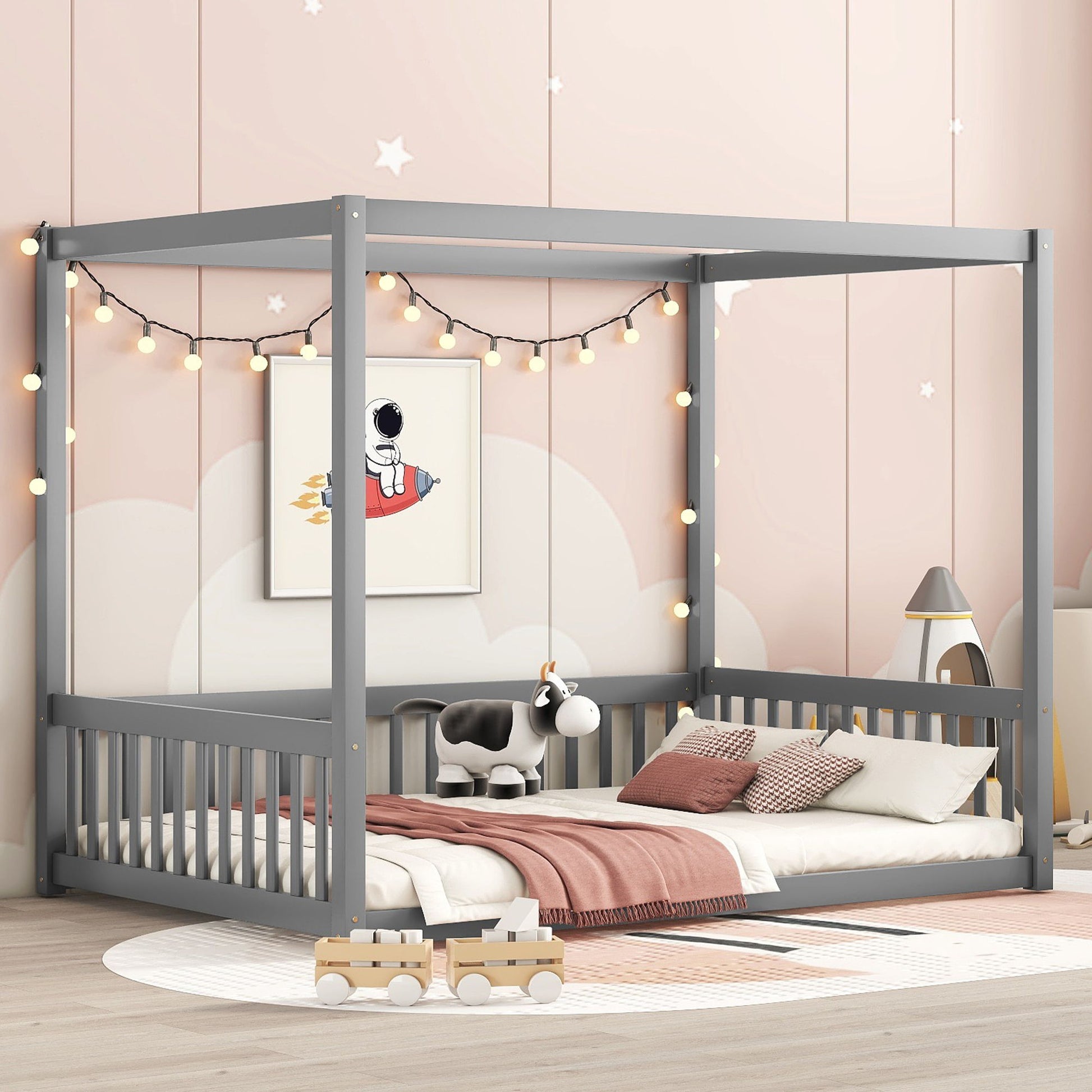 A children's room features a gray Canopy Frame Floor Bed With Fence, adorned with string lights and a cute cow plush sitting among pillows and a pink blanket. The minimalist decor includes pastel tones and wall art of a child on a rocket.