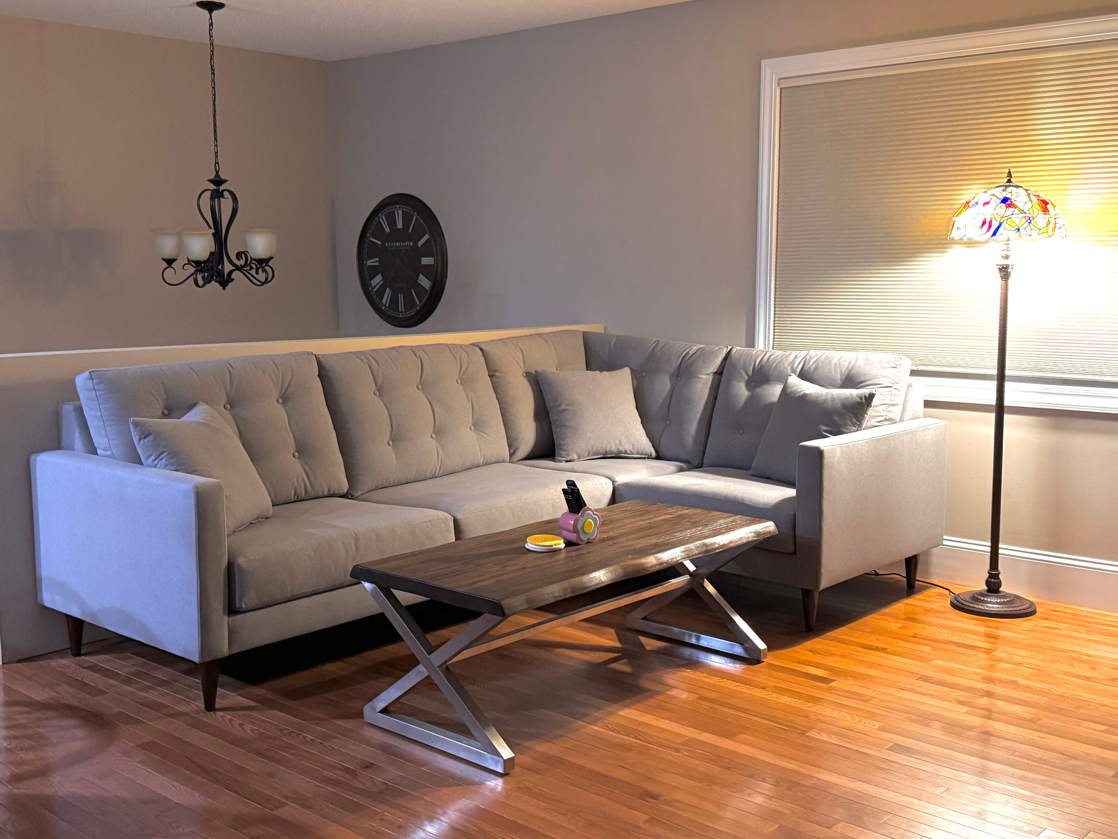 A cozy living room with a gray L-shaped sofa, a wooden coffee table, and a colorful floor lamp. The room features a wall clock and a chandelier, and the wooden floor adds warmth to the space. A window with blinds is in the background.