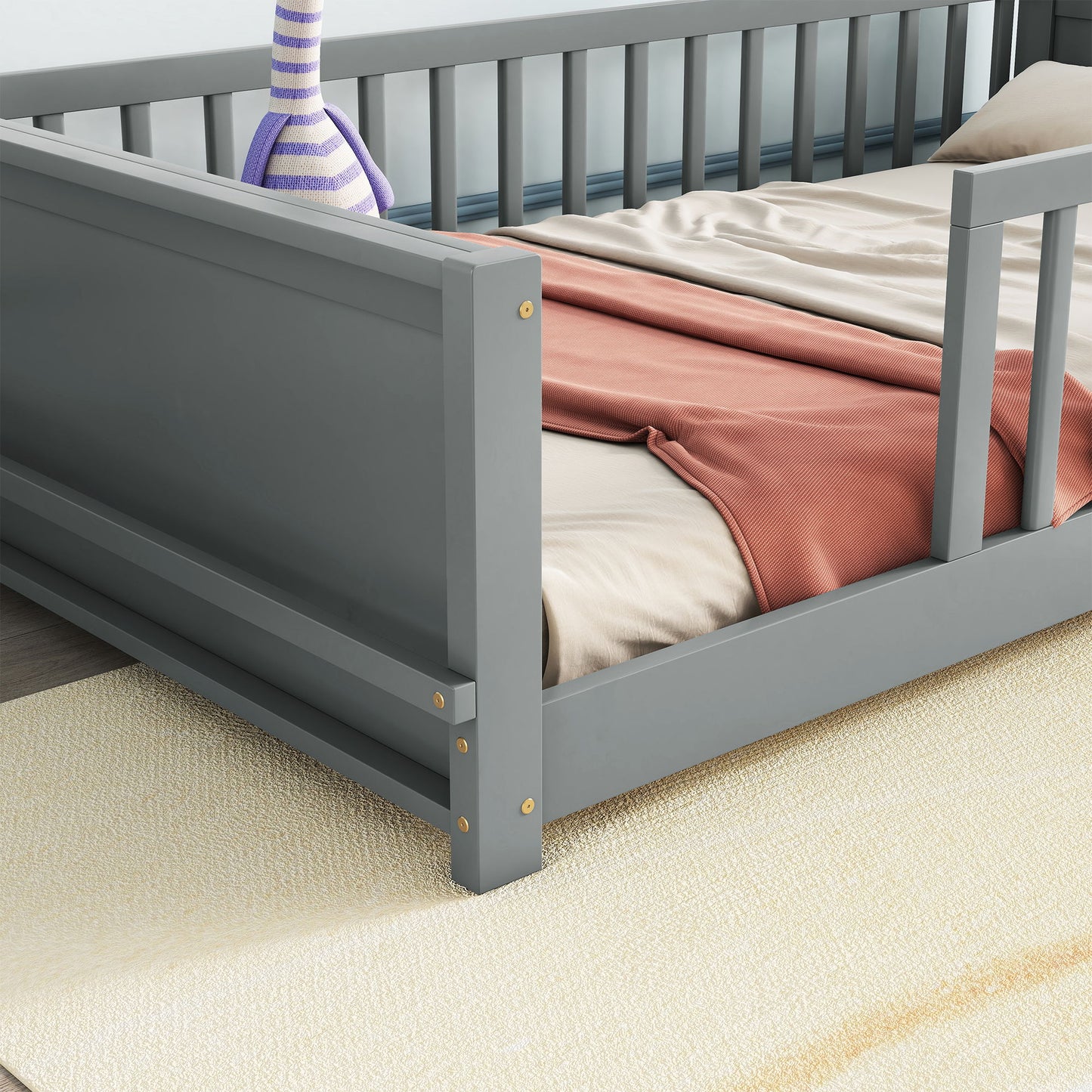 A detailed view of the Floor Platform Bed With Built-In Book Storage Rack, featuring a gray bed frame with a wooden finish, topped by a soft beige sheet and folded coral blanket. It sits on a cream textured carpet, with part of a purple-striped vase visible in the background.