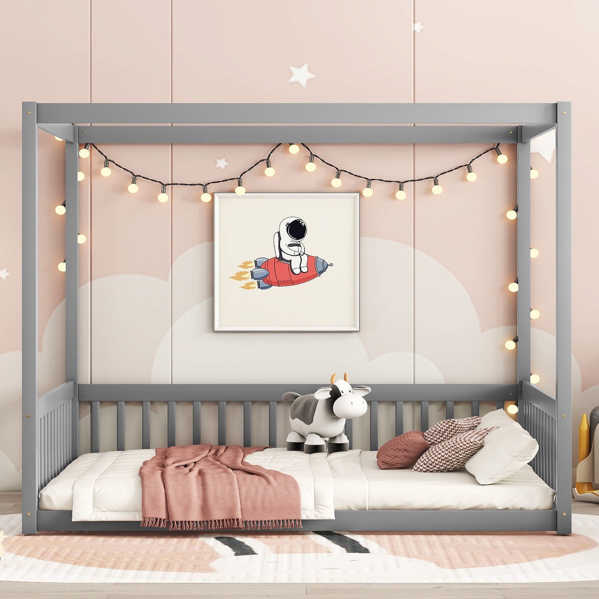 The Canopy Frame Floor Bed with Fence, featuring a gray frame and guardrails, is decorated with string lights. A stuffed cow toy rests on the bed adorned with white and pink bedding. A picture of a cartoon astronaut on a red rocket hangs on the wall.