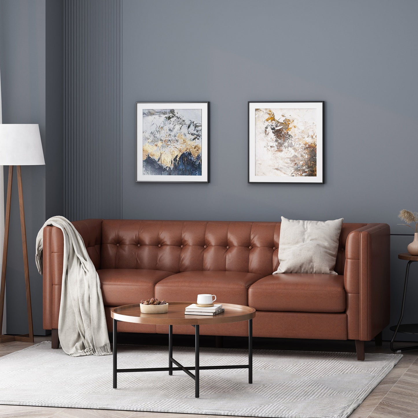 A cozy living room with a Comfy 3 Seat Sofa With Tufted Back, beige pillow, and draped blanket. Two abstract paintings adorn the gray wall above. A modern floor lamp is on the left, while a wooden round coffee table sits in front.