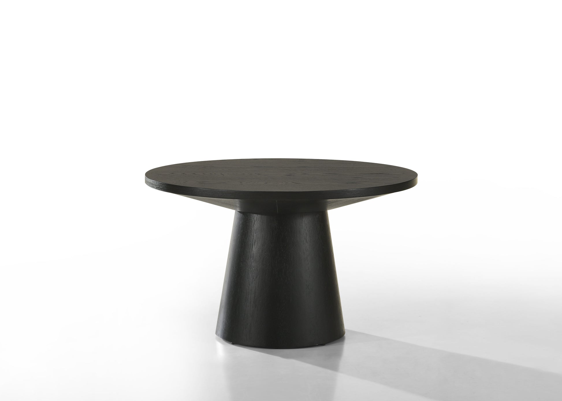 The Jasper - Round Table is a modern piece featuring a round black tabletop with a smooth surface and a wide, tapered cylindrical base, set against a white background.