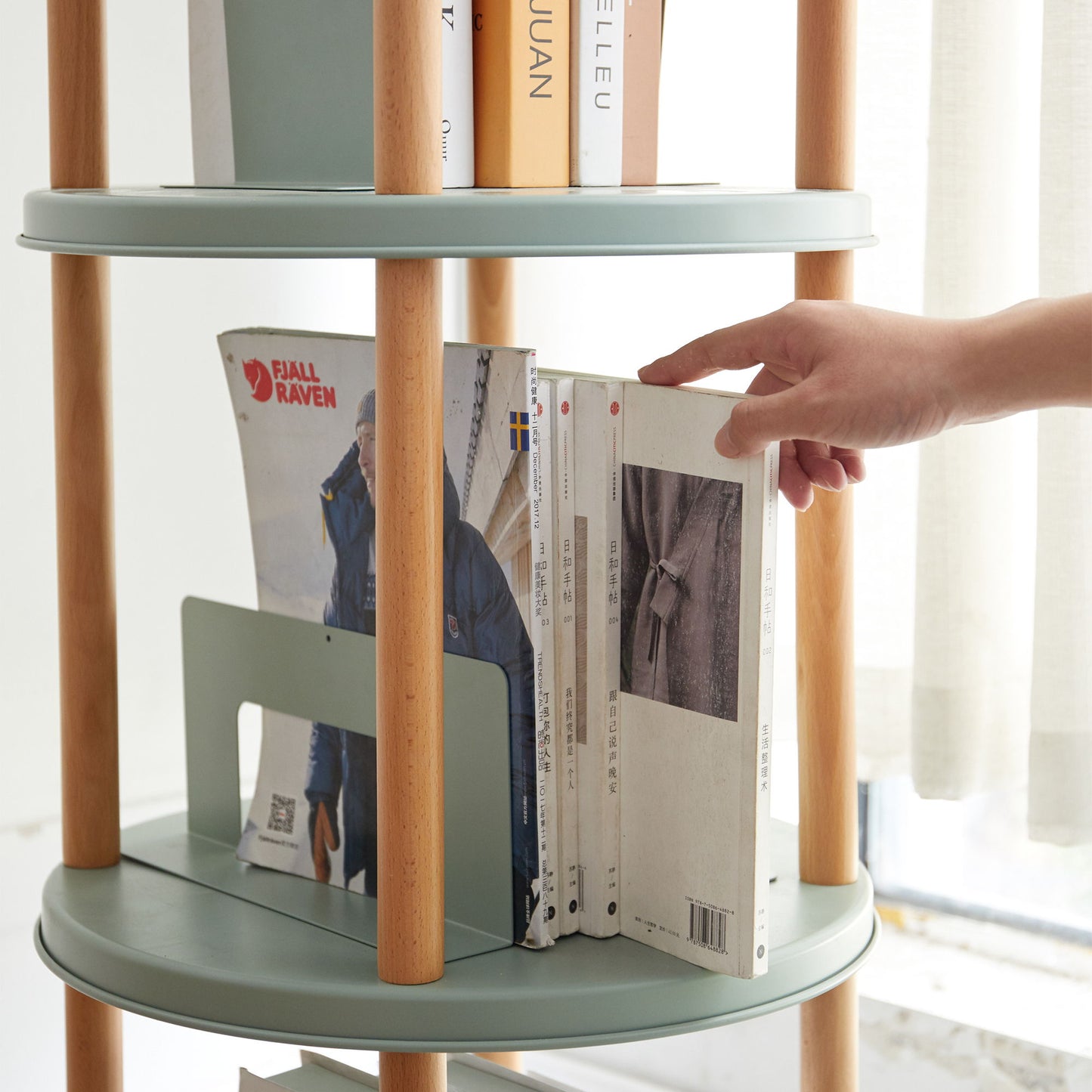 A hand reaches to pick a magazine from a round Storage Shelf, 360° Rotating Bookshelf in light green. The shelf, with natural wooden supports, holds various books and magazines. Sunlight filters through a nearby window covered by light curtains.