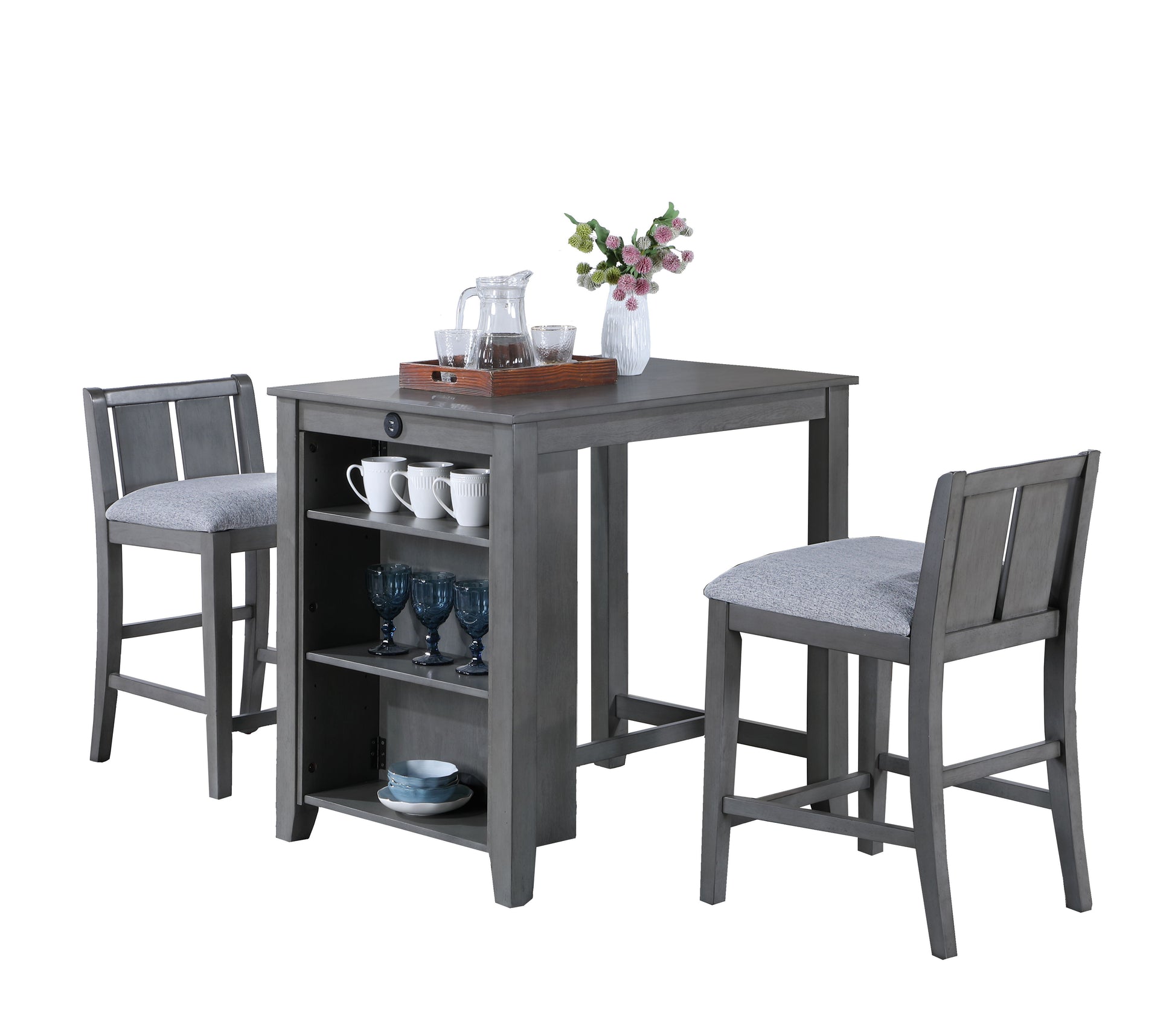 The Graham 3-Piece Set includes a compact gray counter height dining table with built-in shelves for glassware and a tray with a coffee set, flanked by two matching chairs featuring light gray cushioned seats. A vase with pink flowers adorns the tabletop.