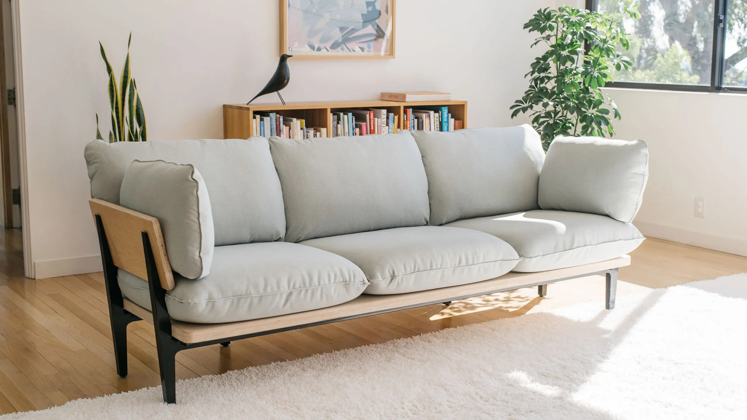 A modern living room features a light blue cushioned sofa with black metal legs on a beige carpet. Behind the sofa, there's a wooden bookshelf filled with books, a plant, and a black bird figurine. Tall green plants and a large window add brightness to the space.