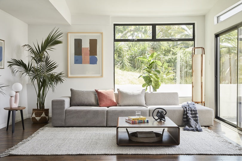 A bright and modern living room features a light gray sofa adorned with a coral pillow and a striped throw. A wooden coffee table holds books and decorative items. Large windows reveal lush greenery outside, while indoor plants and abstract art enhance the aesthetic.