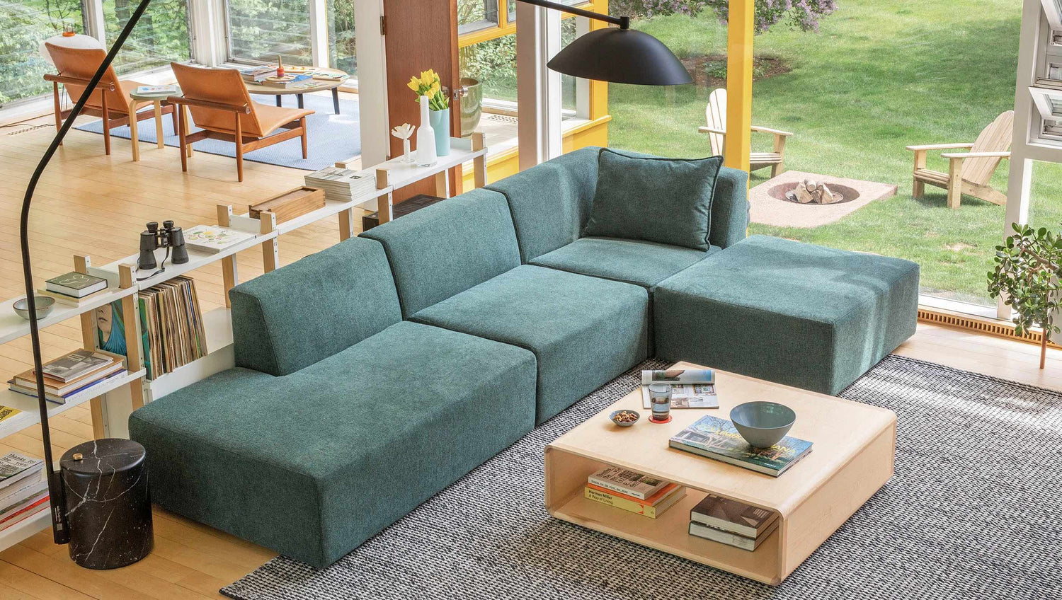 A modern living room with a large teal sectional sofa, a light wood coffee table, and a black floor lamp. The space features abundant natural light from large windows, an open bookshelf, and a cozy seating area in the background by a garden, with wooden furniture.