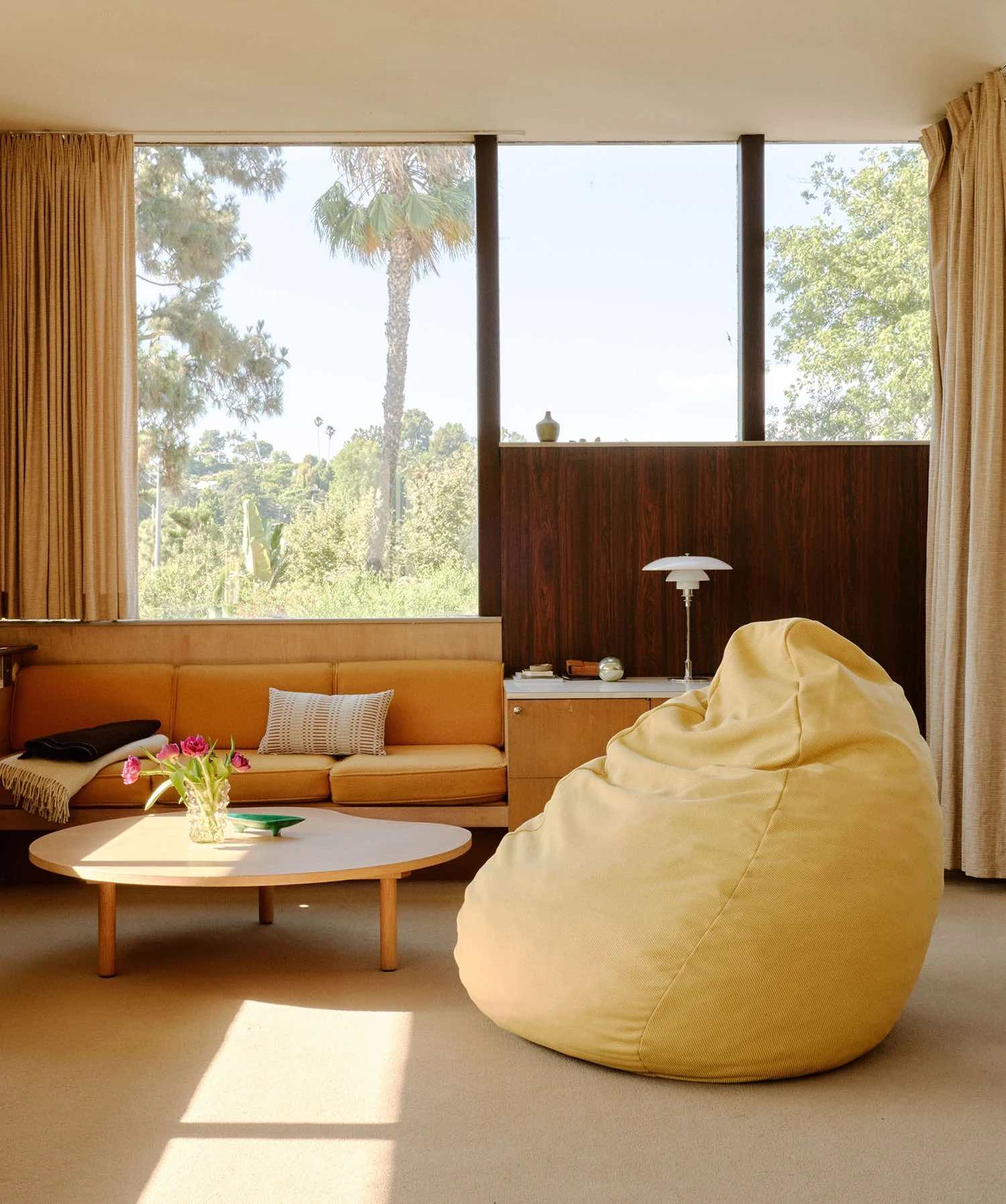A cozy living room with large windows showcasing a scenic outdoor view. The room features a yellow bean bag chair, a round wooden coffee table with a vase of pink flowers, a built-in bench with yellow cushions, and beige curtains.