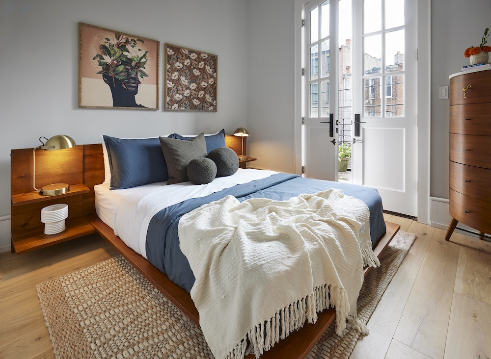 A cozy bedroom featuring a wooden bed with white and navy bedding, adorned with various pillows and a throw blanket. Two bedside tables, each with a lamp, flank the bed. Two framed artworks hang above the bed. French doors lead to an outdoor space.