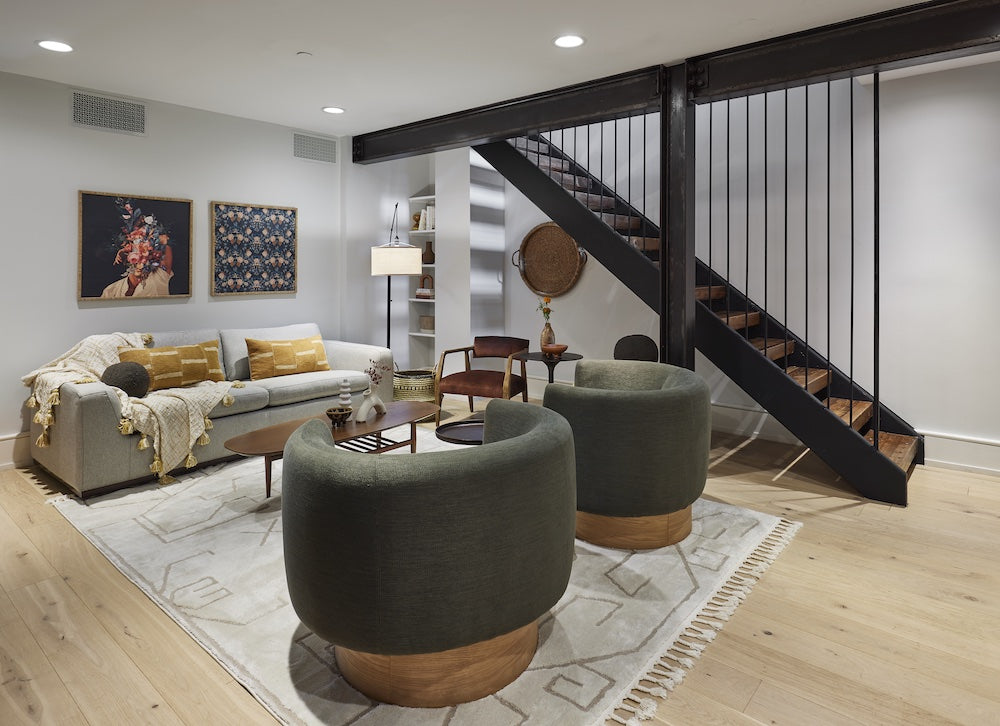 A modern living room with light-colored wood flooring and white walls. It features a gray sofa, two green round swivel chairs, a wooden coffee table, and a staircase with black railings. Artworks, lamps, and decor adorn the space for a cozy, stylish feel.