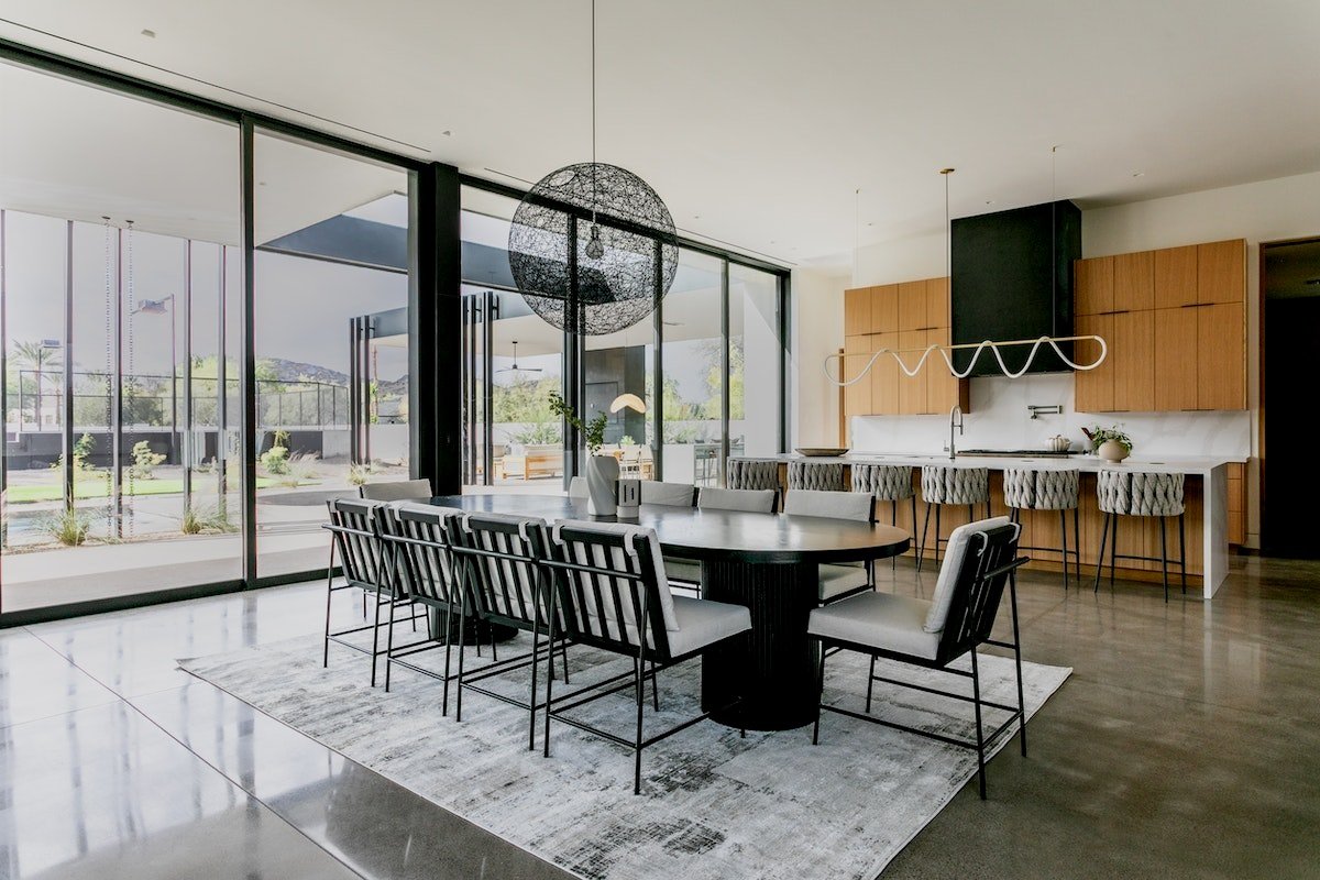 A modern dining area with a long table, black chairs, and an overhead black spherical light fixture. Adjacent is an open kitchen with wooden cabinets, white countertops, and a contemporary light fixture over the kitchen island. Large glass doors open to an outdoor space.