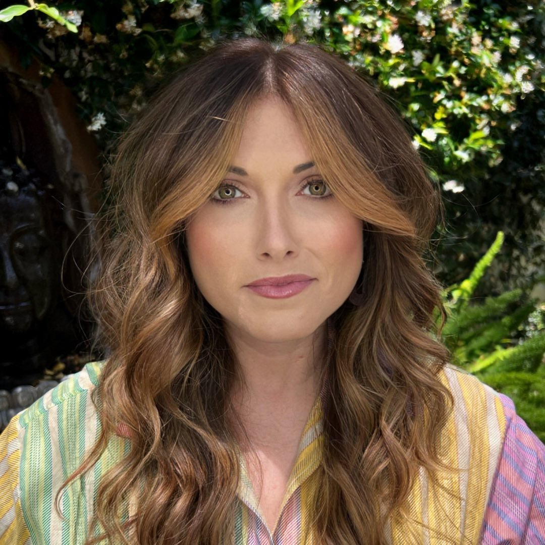 A woman with long, wavy hair stands outdoors in front of greenery and flowers. She wears a colorful, striped shirt and has a calm expression, with subtle makeup enhancing her features.