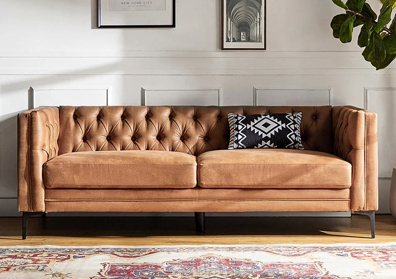 A brown tufted sofa with black legs is placed against a white paneled wall. The sofa has two seat cushions and is adorned with a black and white patterned throw pillow. Above the sofa, two framed pictures are visible. A corner of a patterned rug is on the floor.