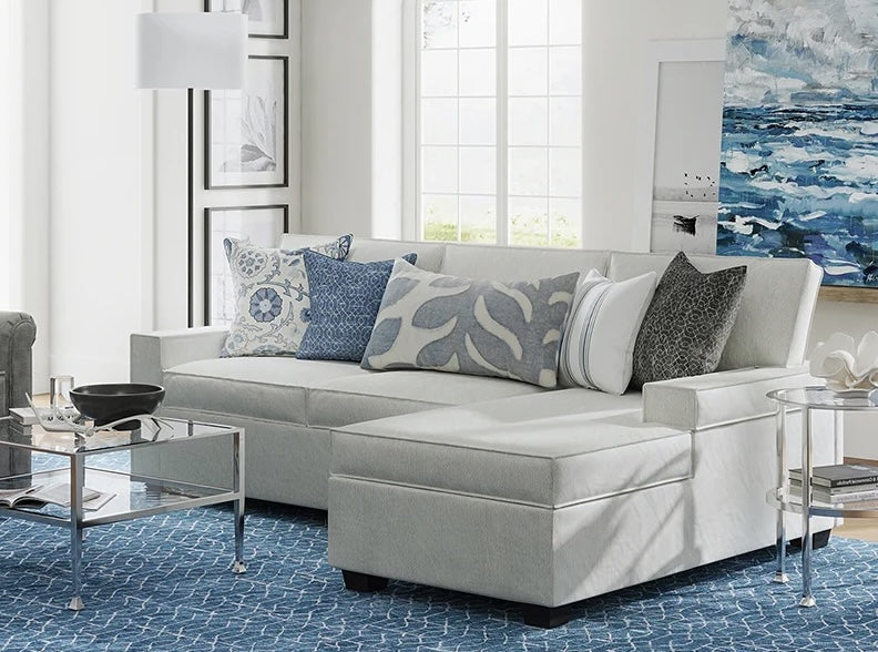 A modern living room featuring a light gray sectional sofa adorned with patterned and solid cushions. A glass coffee table with metal legs holds a black bowl and a book. The room has a blue patterned rug, large windows, and contemporary wall art and decor.